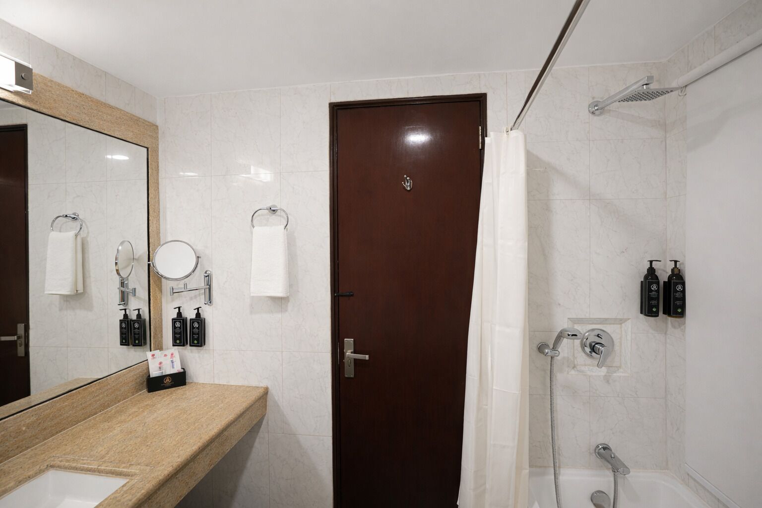 A modern bathroom with a marble vanity, a large mirror, a dark wooden door, and a shower area with a handheld showerhead and two bottles of shampoo.