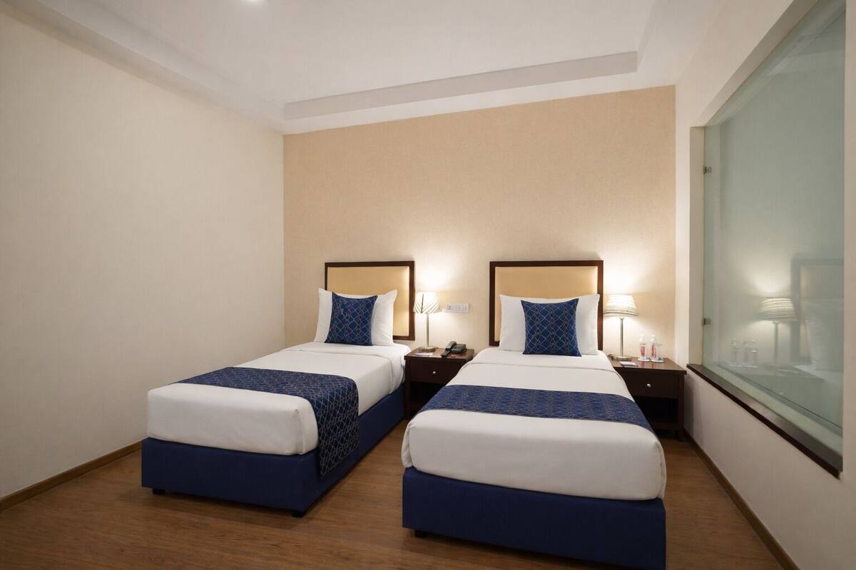 Habitación de hotel con dos camas, cada una con cabecera, mesitas de noche, lámparas y un ventanal.