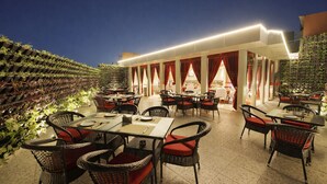 An outdoor dining area with wicker chairs and tables, red curtains, and a wall of plants. - Hotel Amber Palace (Gurugram)