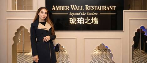 A person standing in front of a sign that reads "AMBER WALL RESTAURANT beyond the borders" with a decorative wall and patterned floor tiles.