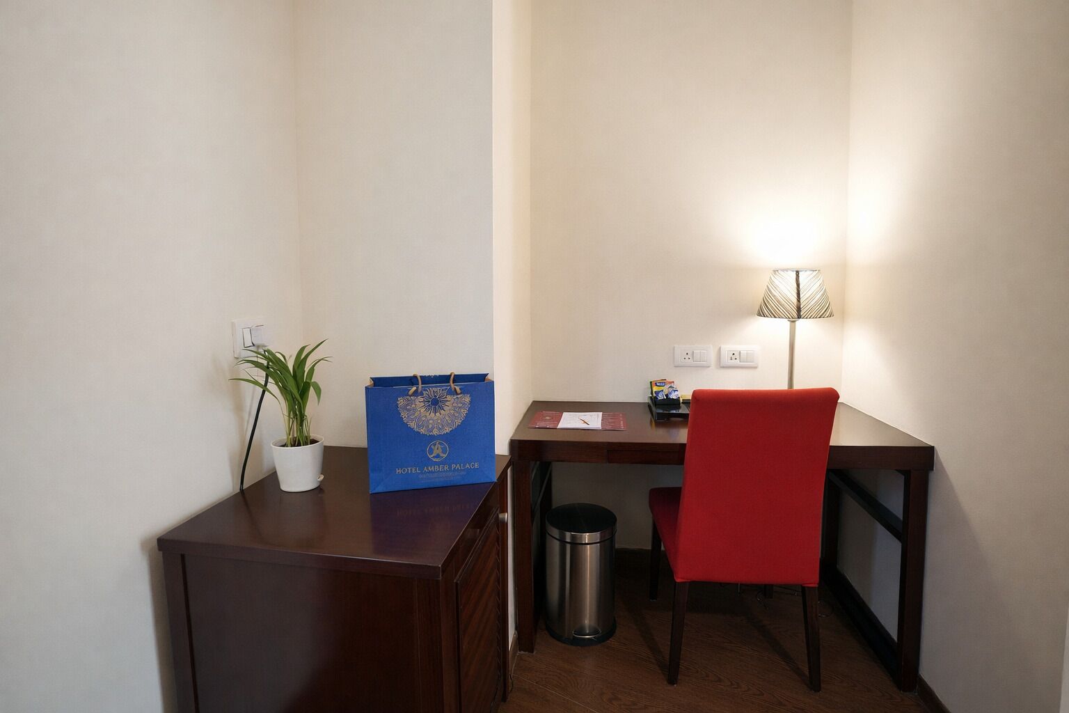 A small, neatly arranged workspace with a desk, chair, lamp, trash bin, and a potted plant.