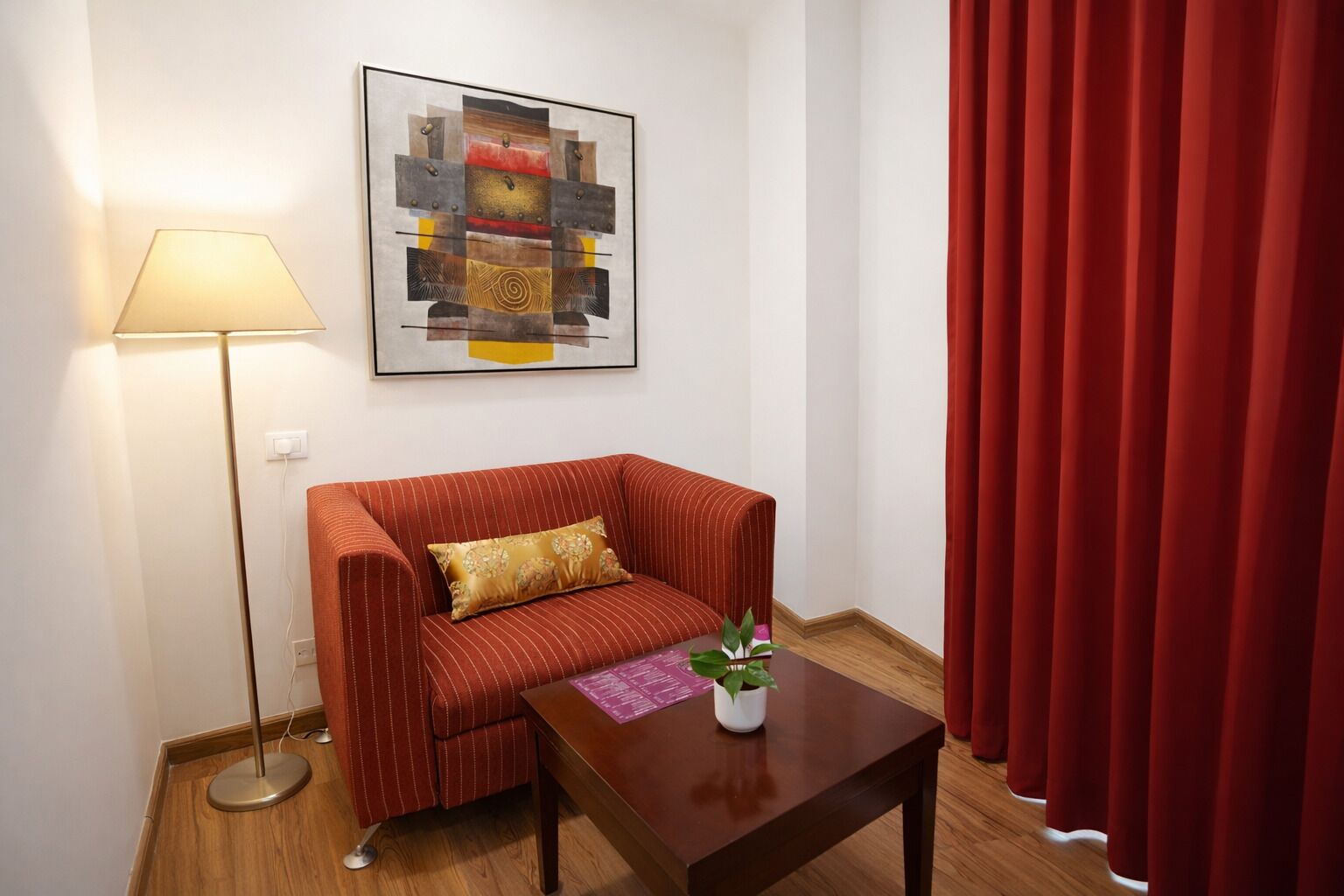 A room with a red-striped sofa, a small wooden table with a plant, a floor lamp, and a red curtain.