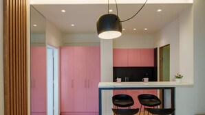 A modern kitchen with pink cabinets and a white countertop. There are three black bar stools under a pendant light. - Irakleio Notos Boutique Apartments 2 (Heraklion)