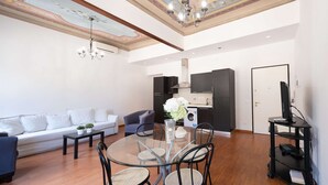 Een moderne woonkamer met een glazen eettafel, zwarte stoelen, een bank, een tv op een meubelstuk en een plafond met een decoratief muurschildering.