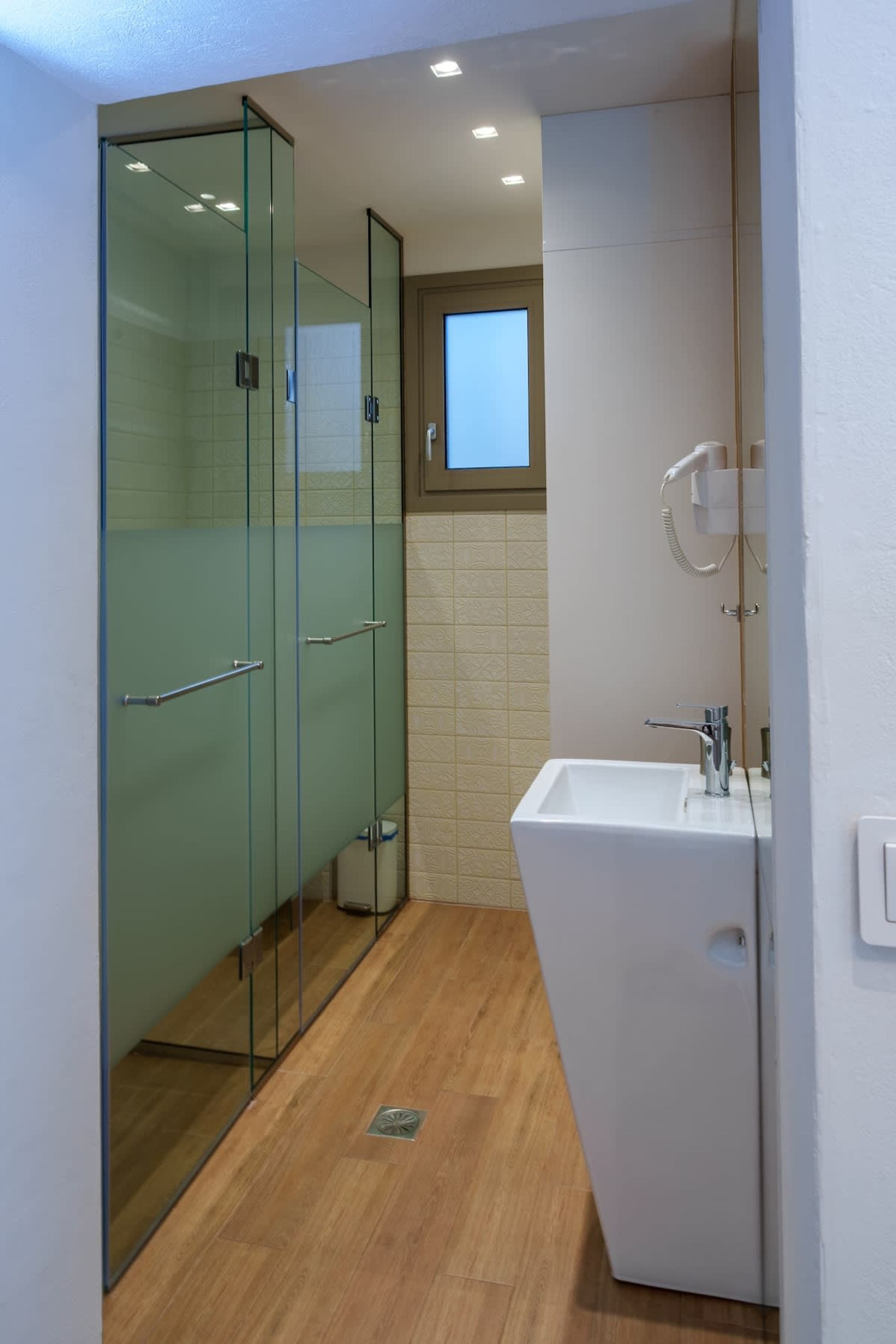 A modern bathroom with a glass shower enclosure, a white sink, and a small window above the mirror.