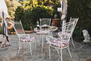 Een terras met witte metalen stoelen en een ronde tafel, omgeven door stenen vloeren en beplanting.