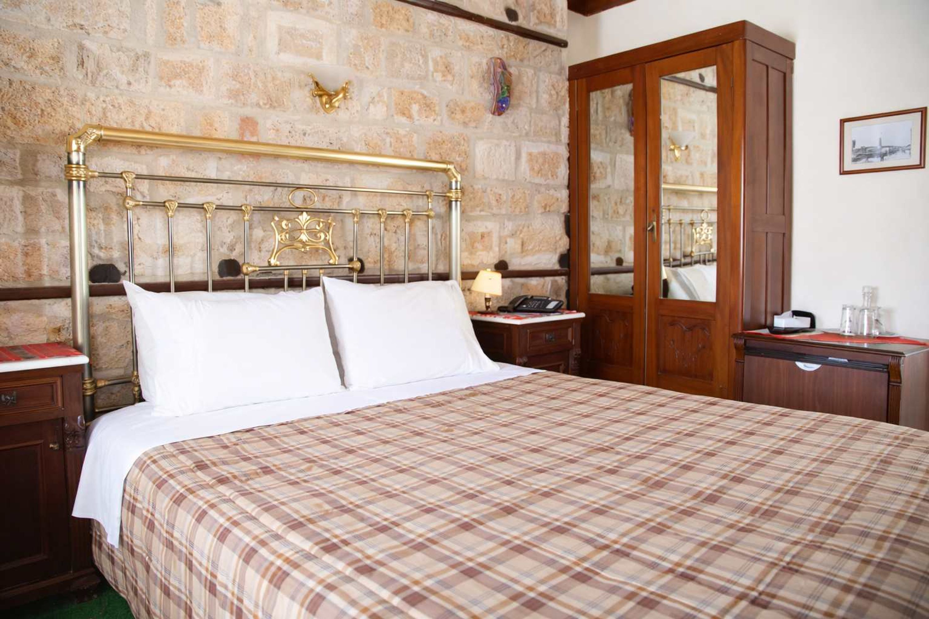 A bed with a plaid bedspread, a wooden nightstand with a lamp, a wooden wardrobe, and a stone wall.