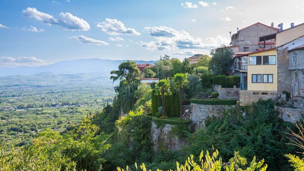 A hillside property with a view of the countryside, featuring a stone retaining wall, a yellow building with a balcony, and lush greenery. - Hagiati Aleksandros - Edessa Cozy Mountain Retreat (Edessa)