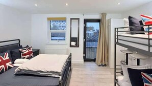 A hotel room with a bunk bed, a single bed, a window with a view of buildings, a door with a curtain, and a radiator. - Entire Flat in Greater London, United Kingdom (London)