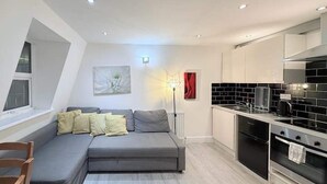 A modern apartment with a grey sectional sofa, a dining area with black tiled backsplash, and a kitchen with white cabinets. - Entire Flat in Greater London, United Kingdom (London)