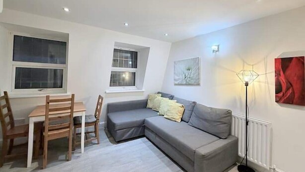 A modern living room with a grey sofa, wooden dining table, and chairs. There are two windows, a radiator, and a floor lamp. - Entire Flat in Greater London, United Kingdom (London)