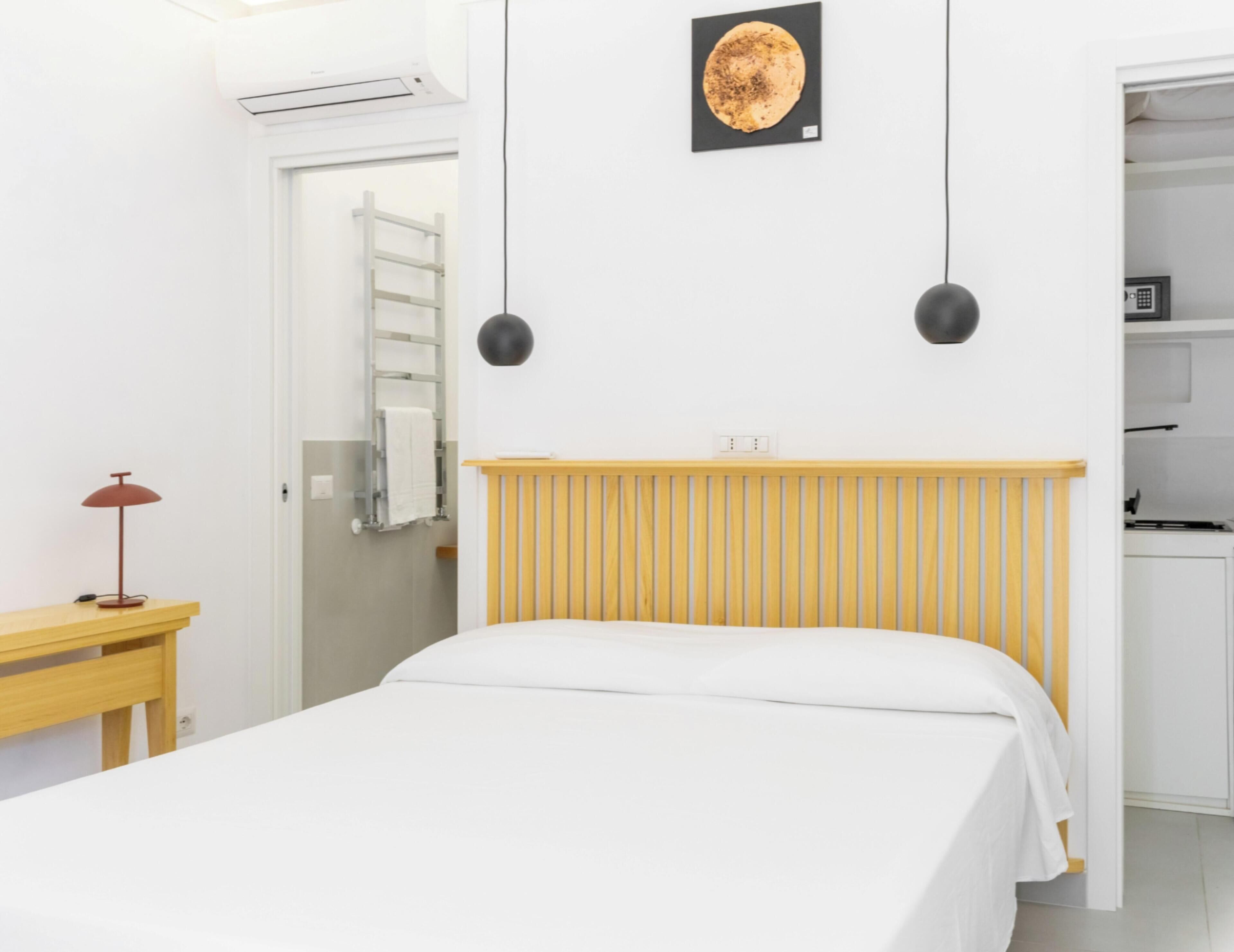 A bedroom with a bed, a wooden bedside table, a red lamp, a white wall, a black pendant light, a white door, and a small kitchen area.