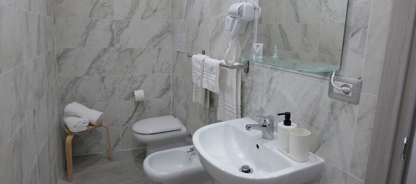 Une salle de bain avec un mur en marbre, des toilettes, un bidet, un lavabo avec un miroir et un sèche-cheveux fixé au mur.