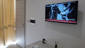 Une salle de bain moderne équipée d’un téléviseur mural diffusant une chaîne d’information, d’un porte-serviettes et d’une petite table sur laquelle sont posés une bougie et un verre.