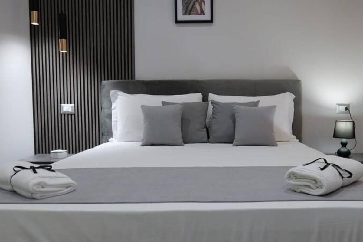 A neatly made bed with pillows and a blanket, a bedside table with a lamp, and a wall-mounted light fixture.
