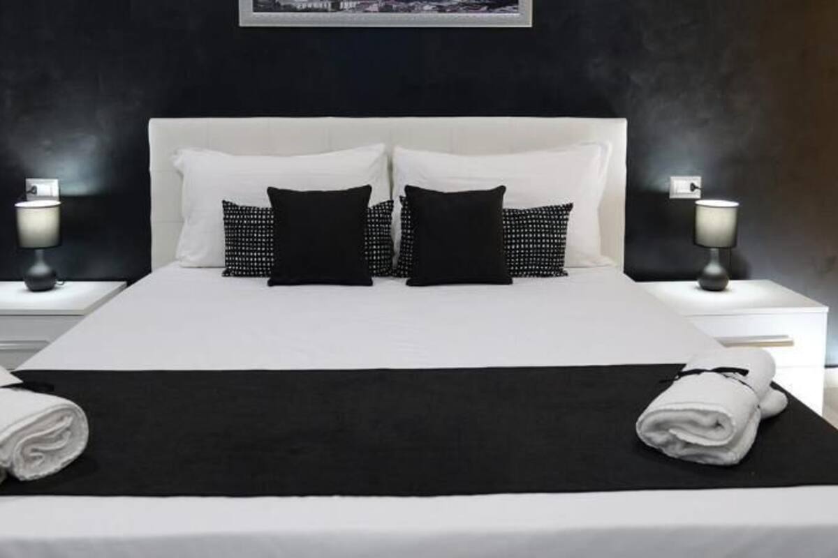 A neatly made bed with white and black bedding, two bedside tables with lamps, and a dark wall with a framed picture.