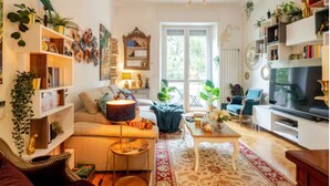 Una sala de estar acogedora con un sofá, una mesa de centro y un televisor. Hay plantas, estanterías y una ventana con persianas.