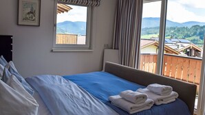 Een slaapkamer met een bed, een raam met gordijnen, een ingelijst schilderij aan de muur en uitzicht op bergen en gebouwen.