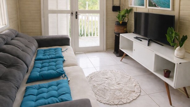 Un salon avec un canapé gris, un téléviseur posé sur un meuble blanc et une porte donnant sur un balcon.