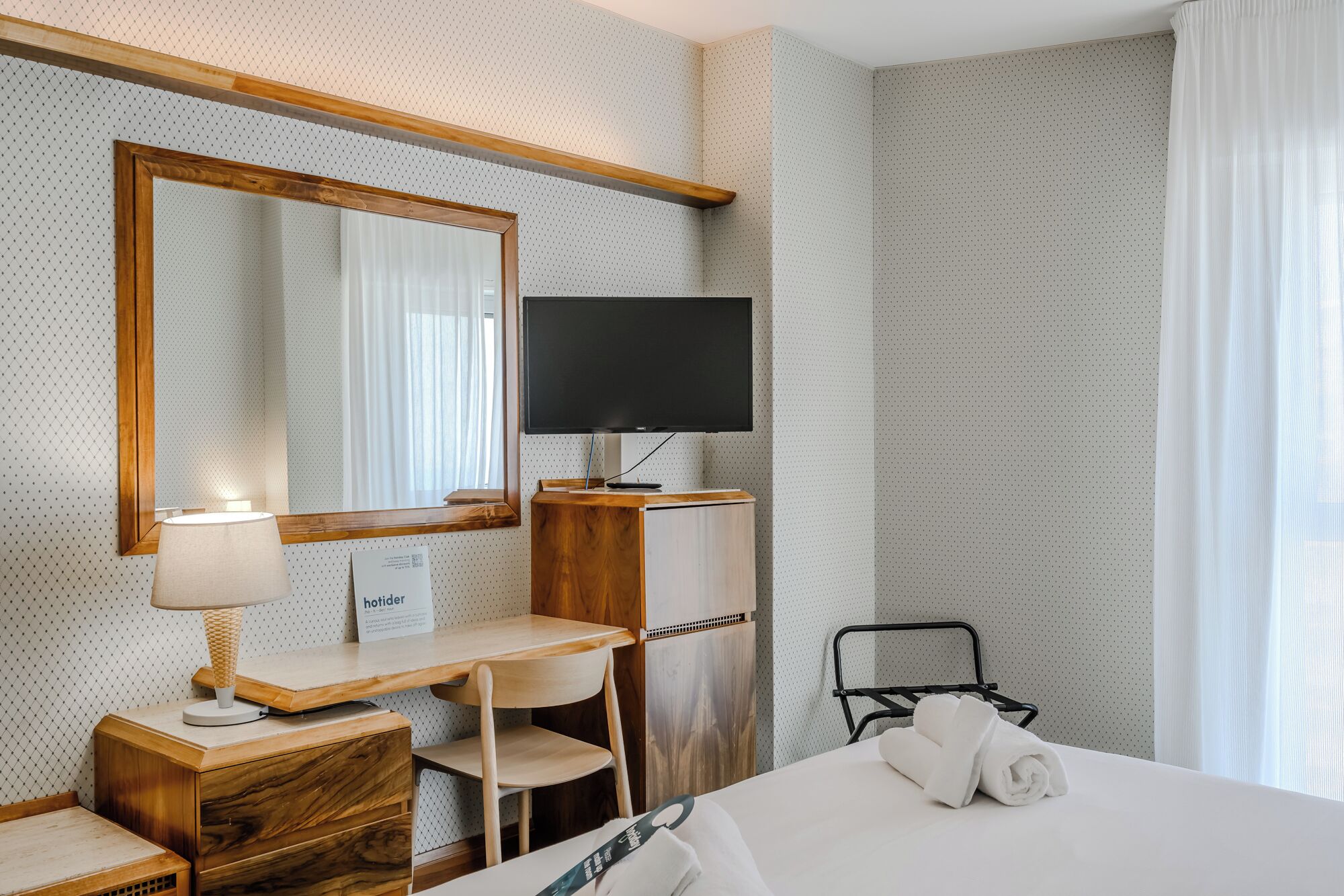 A hotel room with a bed, a desk with a lamp, a TV, a chair, and a bathroom area.