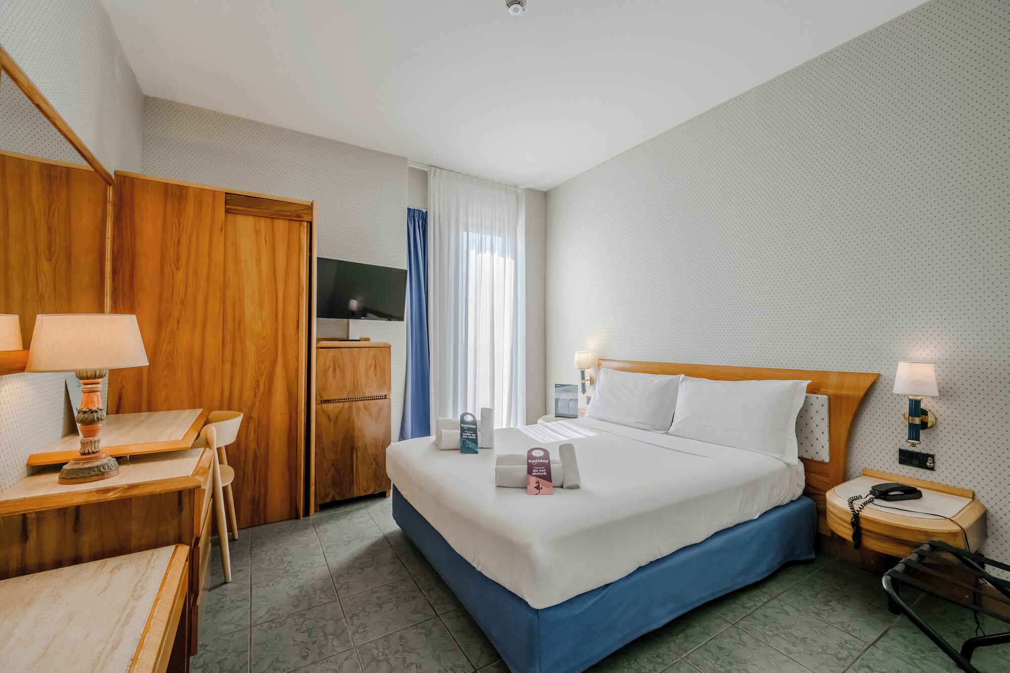 A hotel room with a bed, desk, chair, TV, and a minibar.