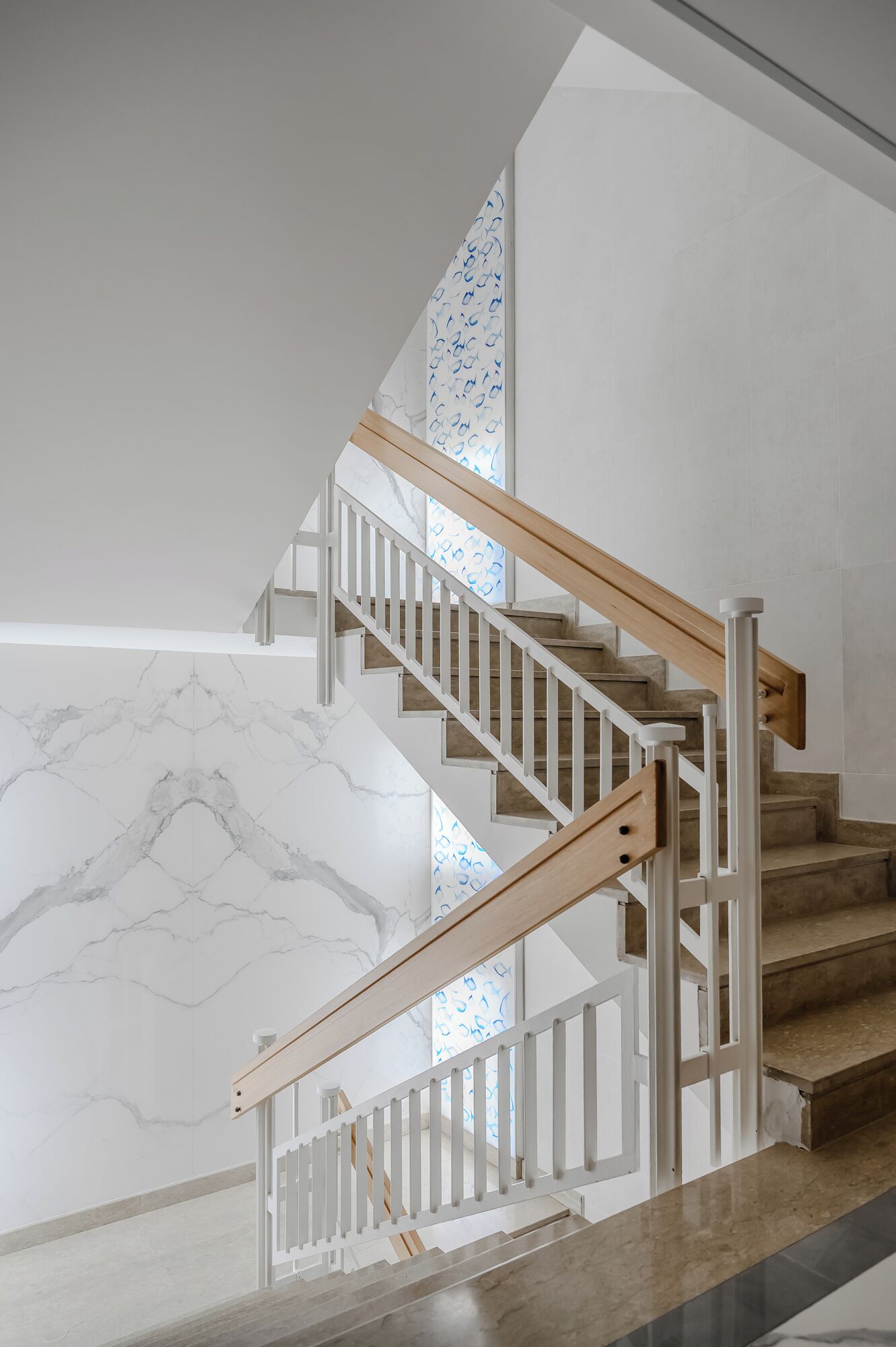 Un escalier avec des rampes blanches et des marches en bois, menant à un palier doté d’un mur en marbre.