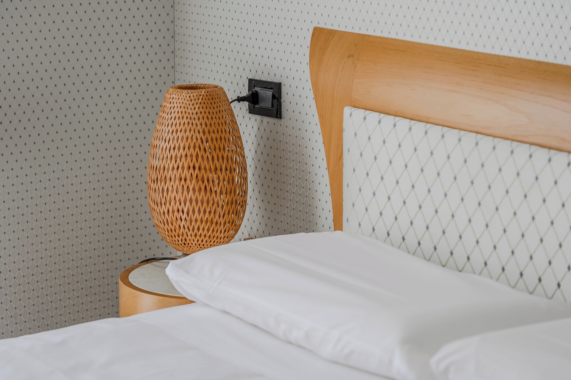 Un lit avec du linge de lit blanc, une tête de lit en bois et une lampe en osier sur une table de chevet.