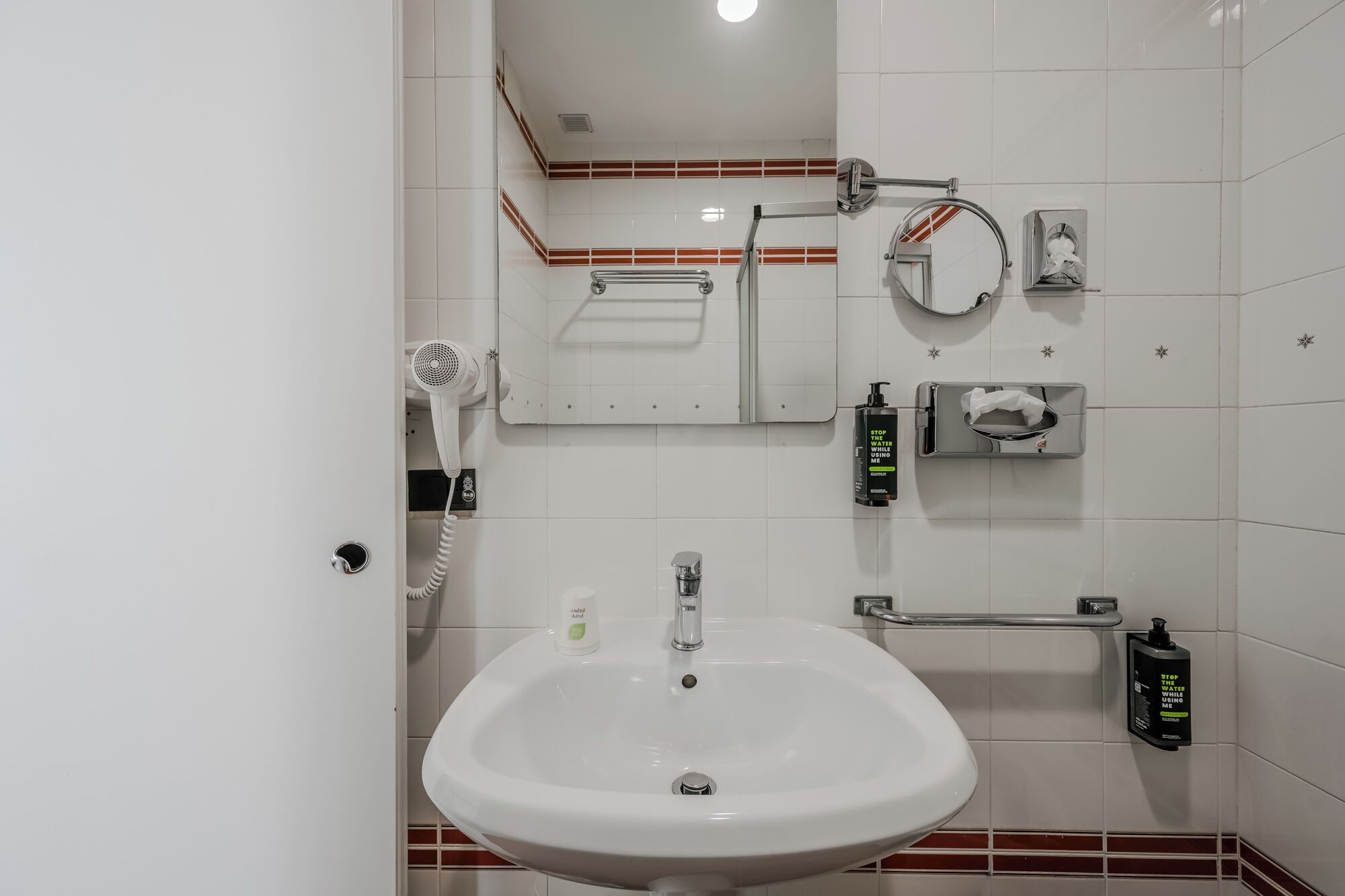 Un baño con lavamanos blanco, secador de pelo, espejo y dispensadores de gel antibacterial.