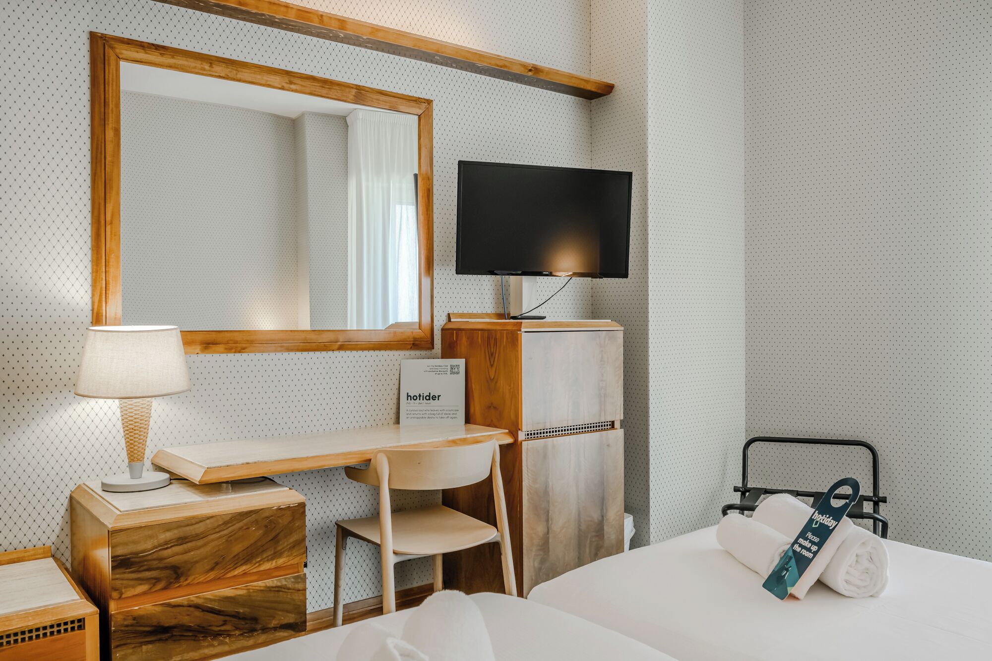 A hotel room with a bed, a desk, a chair, a TV, a mirror, and a towel rack.