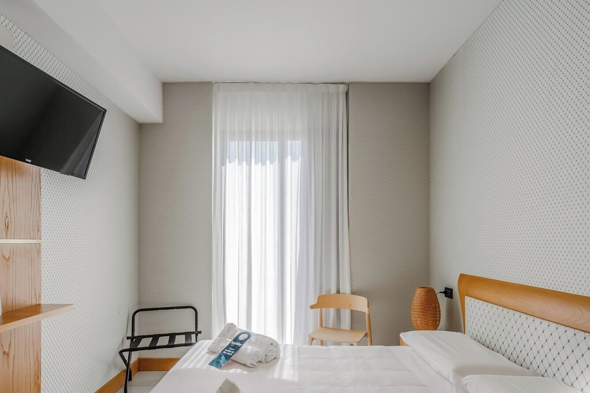 A hotel room with a bed, a chair, a TV mounted on the wall, and a window with curtains.