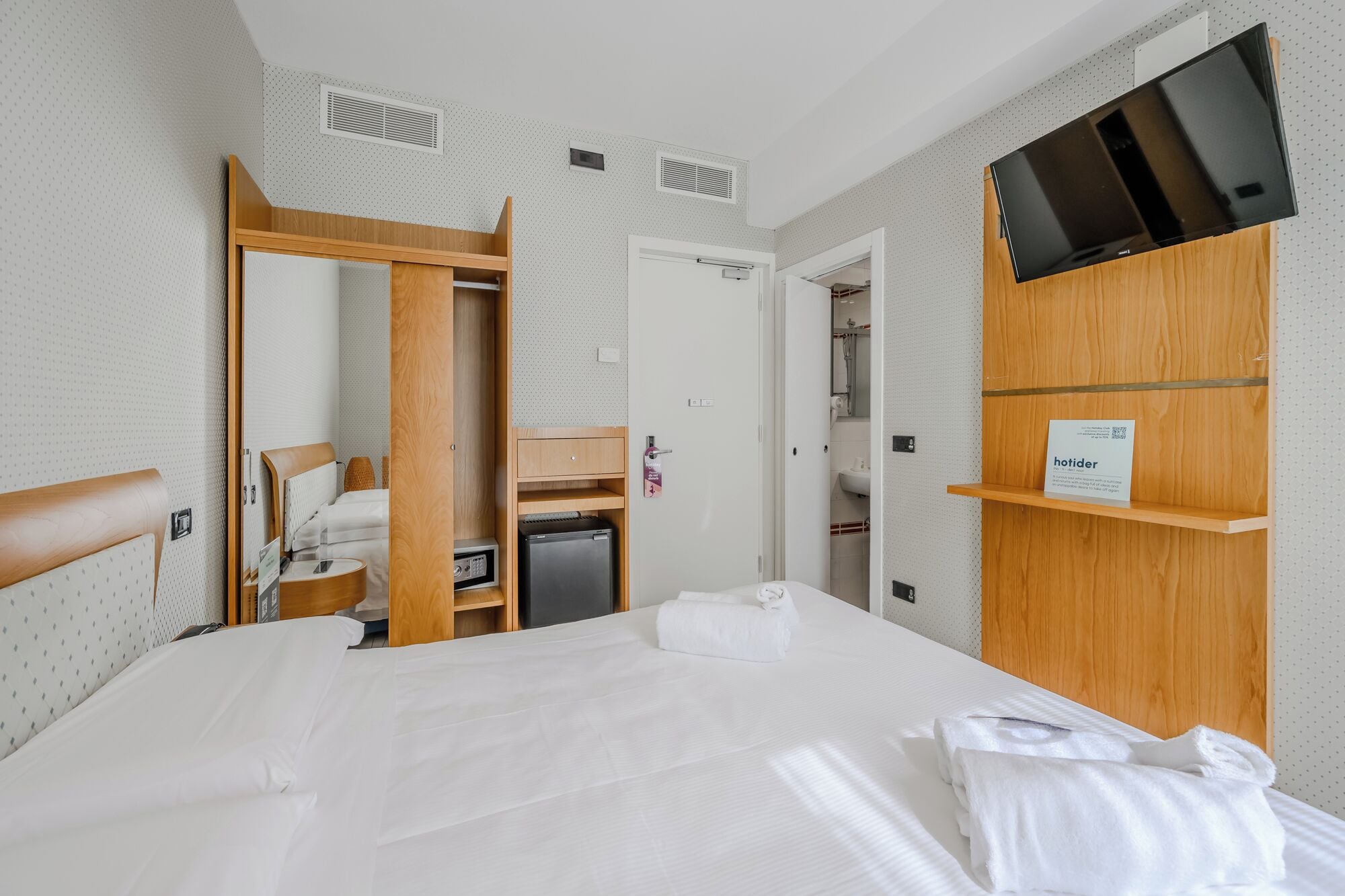 A hotel room with a bed, a wooden wardrobe, a television, a mini-fridge, and a bathroom visible through an open door.