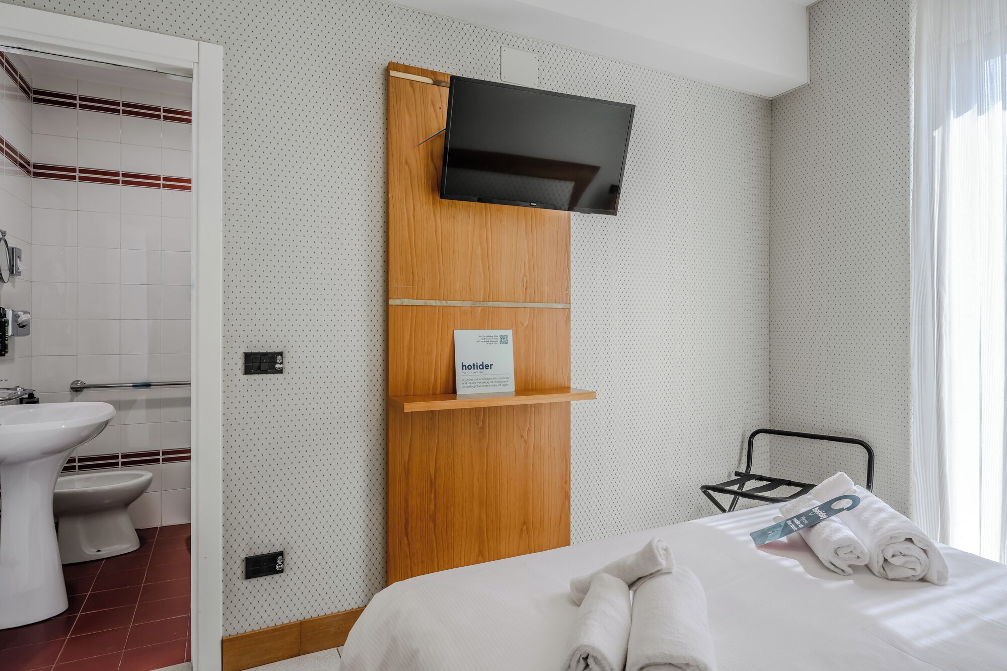 Quarto de hotel com prateleira de madeira, televisão, cama com toalhas, pia e sanita.