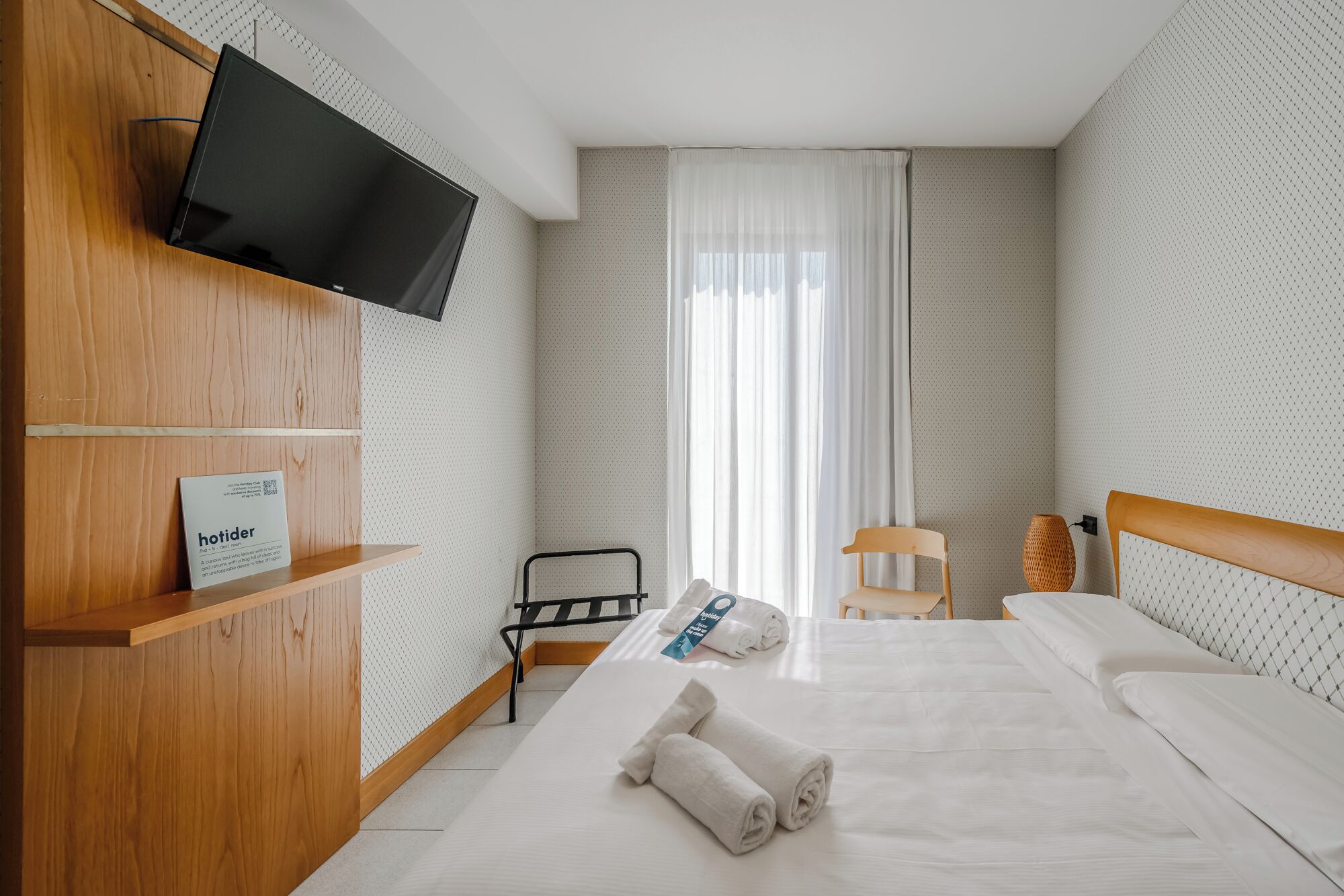 A hotel room with a bed, a TV mounted on a wooden wall, a chair, a small table, and a window with curtains.