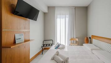 A hotel room with a bed, a TV mounted on a wooden wall, a chair, a small table, and a window with curtains.