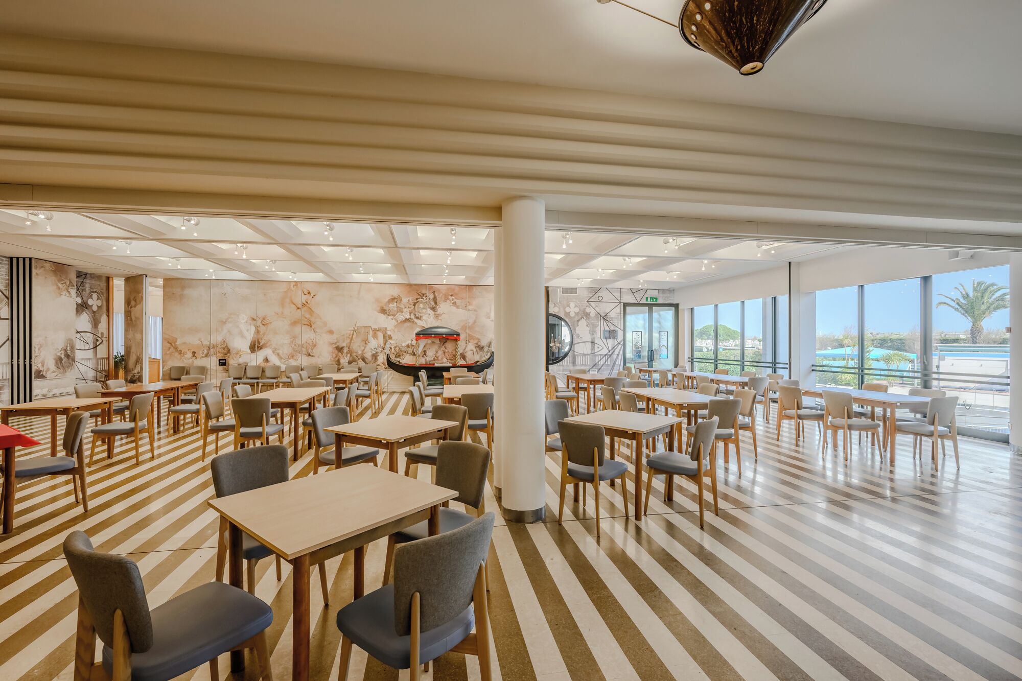 A spacious dining area with striped flooring, multiple tables and chairs, and large windows offering a view of the outdoors.