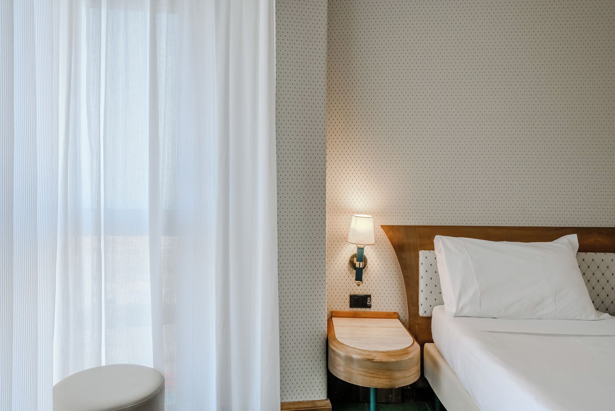 A hotel room with a bed, a nightstand with a lamp, a wooden side table, and a window with sheer curtains.