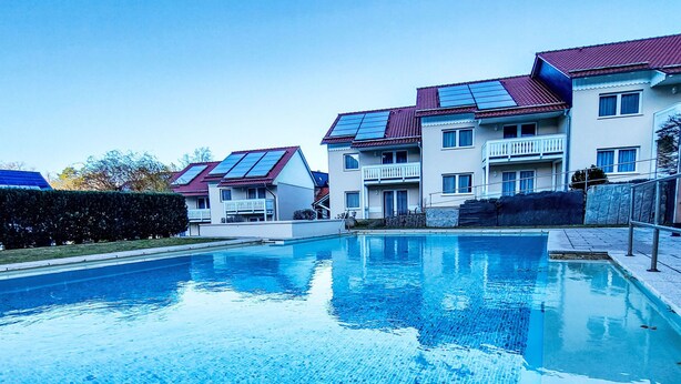 En swimmingpool med fliseterrasse, omgivet af boligbygninger med solpaneler på taget.