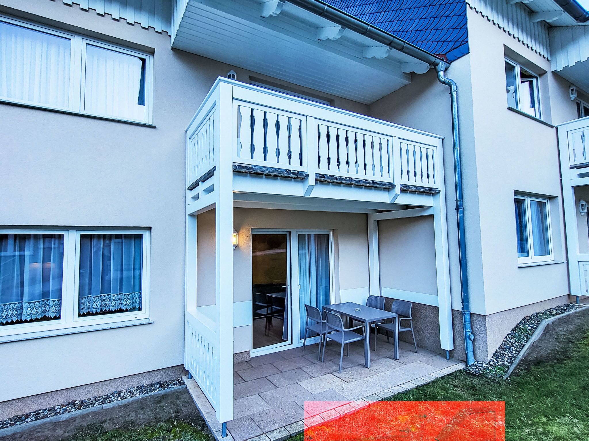 A two-story residential building with a balcony, a patio with a table and chairs, and a small garden area.
