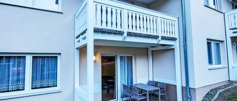 A two-story residential building with a balcony, a patio with a table and chairs, and a small garden area.