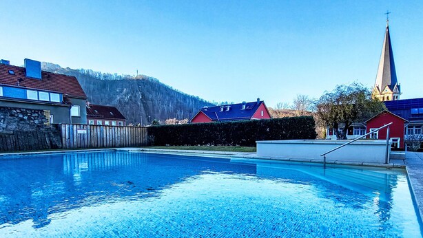 A swimming pool with a clear blue water surface, surrounded by houses and a church with a tall steeple. - Hotelpark-Bodetal by Interhome (Thale)