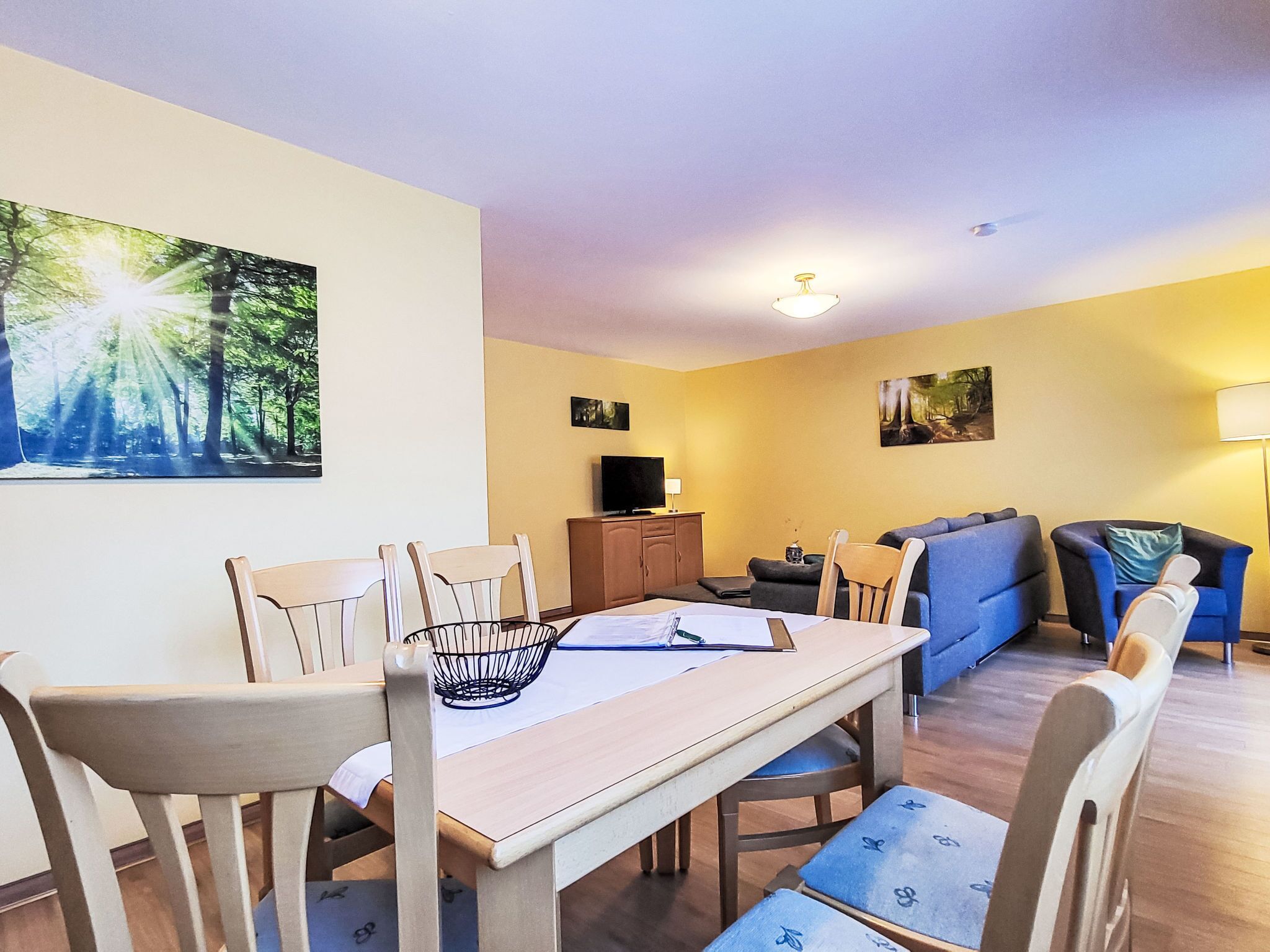 A dining area with a table, chairs, and a television. There is a sofa and a lamp in the living area.