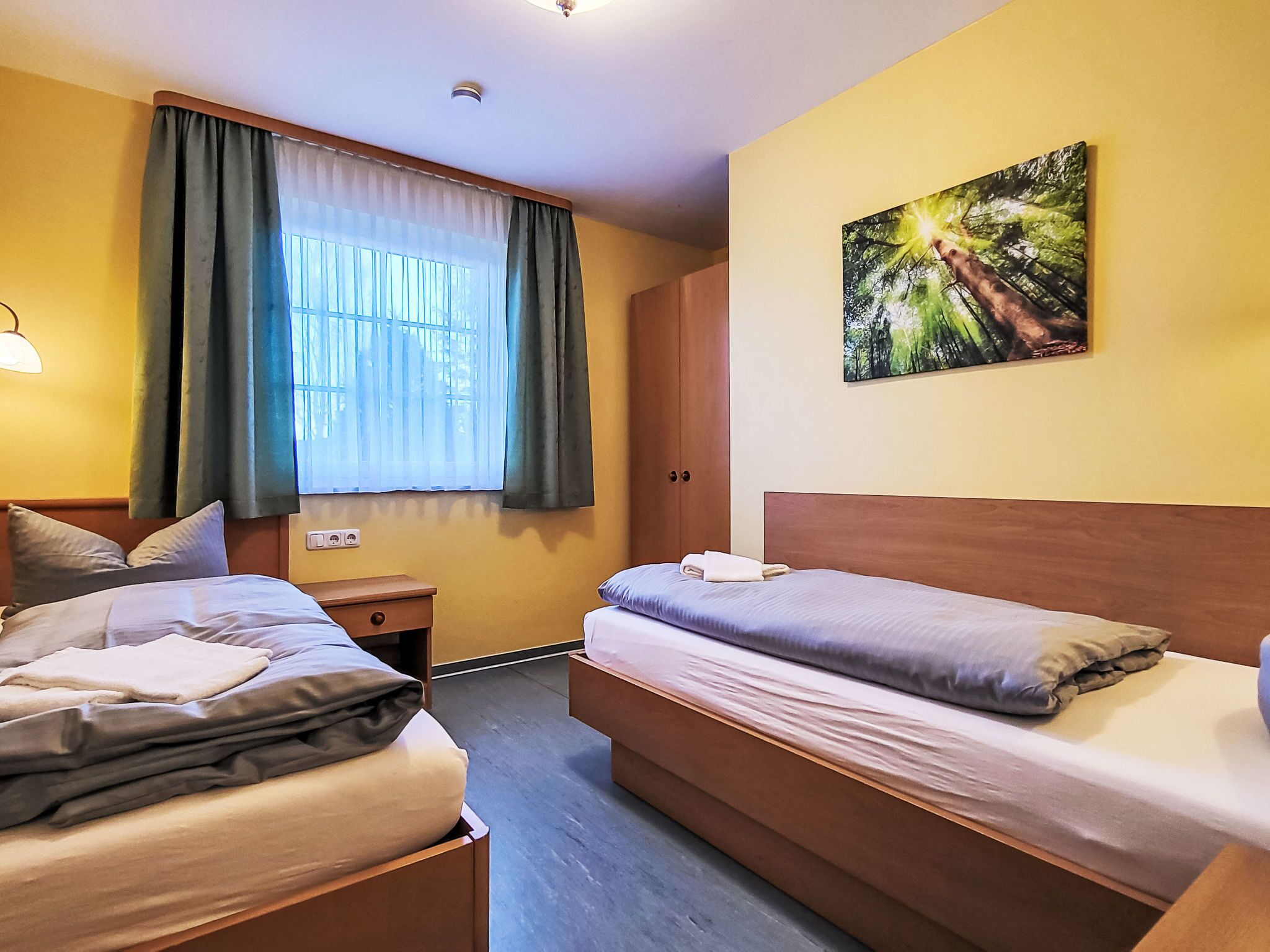 Habitación de hotel con dos camas, una ventana con cortinas, una mesita de noche, un cuadro en la pared y una lámpara.