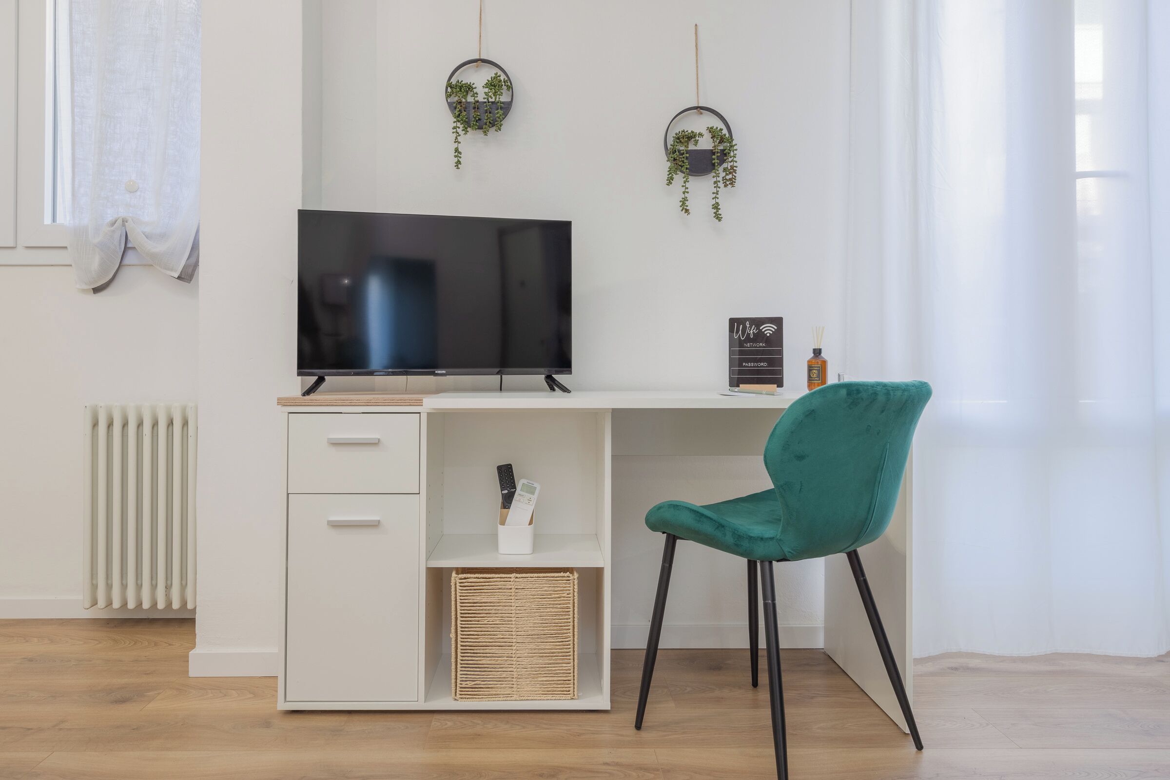 Ein modernes Zimmer mit weißem Schreibtisch, schwarzem Fernseher, blaugrünem Sessel und zwei hängenden Pflanzentöpfen.