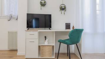 Ein modernes Zimmer mit weißem Schreibtisch, schwarzem Fernseher, blaugrünem Sessel und zwei hängenden Pflanzentöpfen.