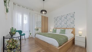 Een moderne slaapkamer met een houten bed, groen beddengoed, een klein nachtkastje, een stoel, een bureau met een plant en een grote spiegel.