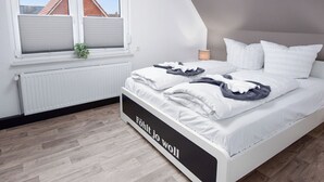 Een moderne slaapkamer met een schuine plafond, een groot raam met zonwering, een radiator, een bedlampje en netjes opgevouwen kleding op het bed.