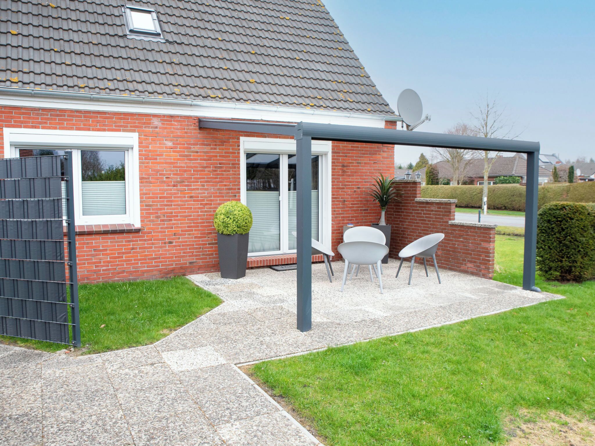 Ein Haus mit Ziegelaußenwand, grauem Dach, Satellitenschüssel, einer Terrasse mit grauer Umzäunung und einem kleinen Garten.
