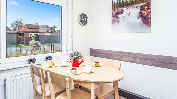 Een eetruimte met een ronde tafel, stoelen, een raam met uitzicht op een huis en een standbeeld, een klok en een lijst met een foto van een waterval.