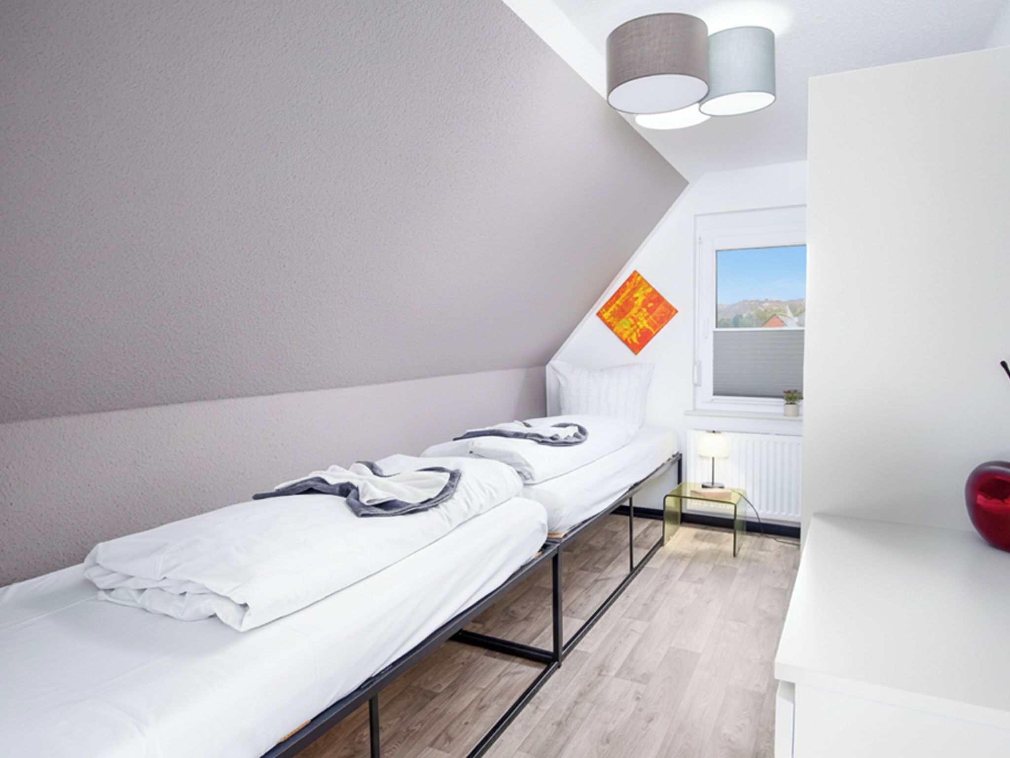 Un lit simple avec un cadre en métal, une table de chevet, une lampe et une fenêtre donnant sur l’extérieur.
