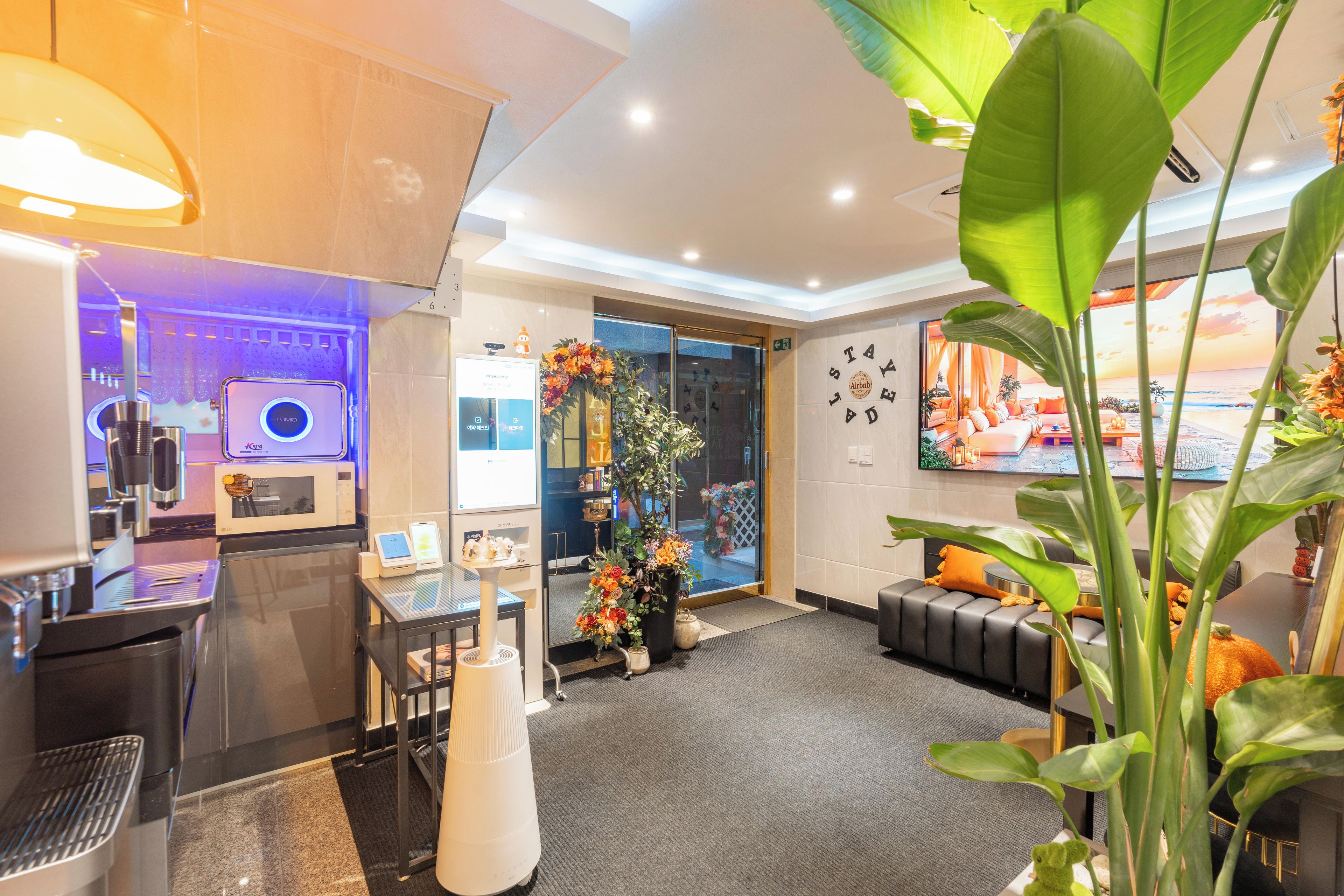 Un hall d’hôtel moderne comprenant un comptoir d’accueil, un espace salon avec un canapé et des fauteuils, une grande plante et un écran mural diffusant une vue panoramique.